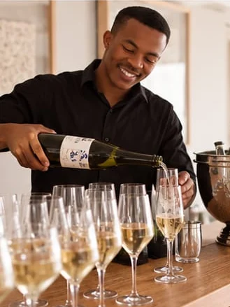 A bartender in a black shirt pours sparkling wine into a row of flutes on a bar counter.